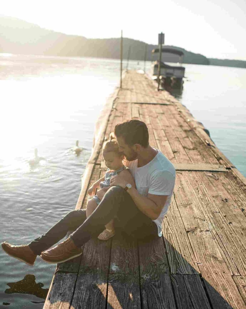 Bild eines Mannes, der an einem Steig eines Sees sitzt und ein Kleinkind im Arm hält